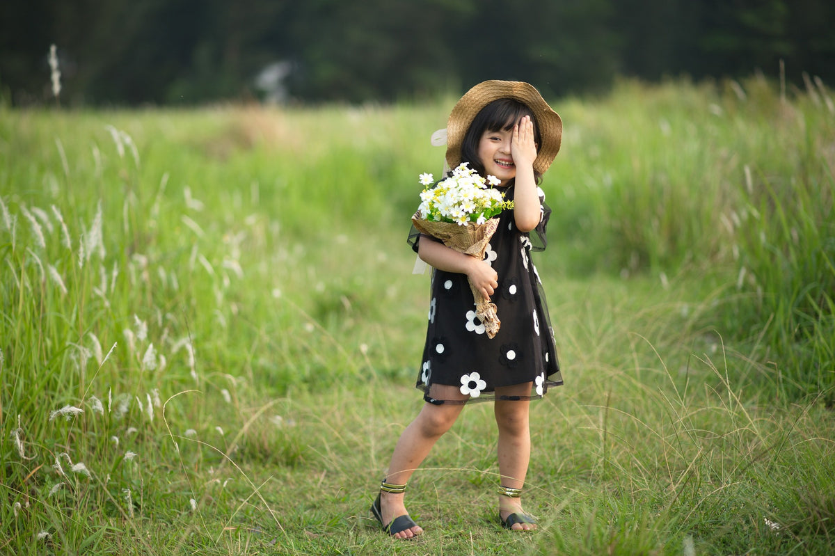 4-year-old girl in a black and white floral dress holds a bouquet of white flowers and playfully covers one eye. At 4 years of age, children should be strengthening some essential life skills such as communication, self-advocacy, and self-care.